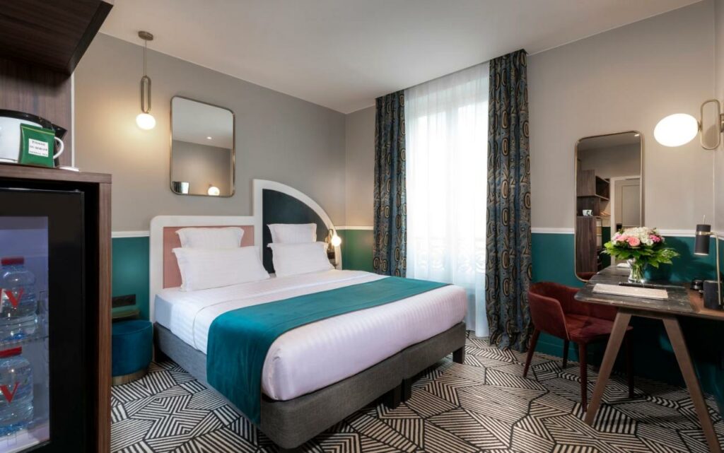 Quarto do Hotel Elysa-Luxembourg, um dos hotéis no Quartier Latin em Paris. Uma cama de casal está ao lado esquerdo da imagem com mesinhas de cabeceira com luminárias dos dois lados, bem como um espelho na parede acima. Na lateral direita da cama fica a janela com cortinas, e logo ao lado há uma mesa de trabalho com cadeira, abajur e vaso de flores. Ao lado esquerdo da cama está o frigobar.
