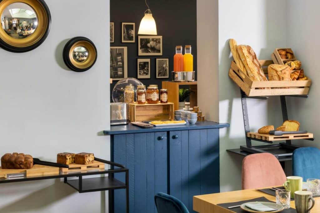 Salão de café da manhã do Hôtel Hélios Opéra com mesinhas postas e pães, sucos e geleias na bancada.