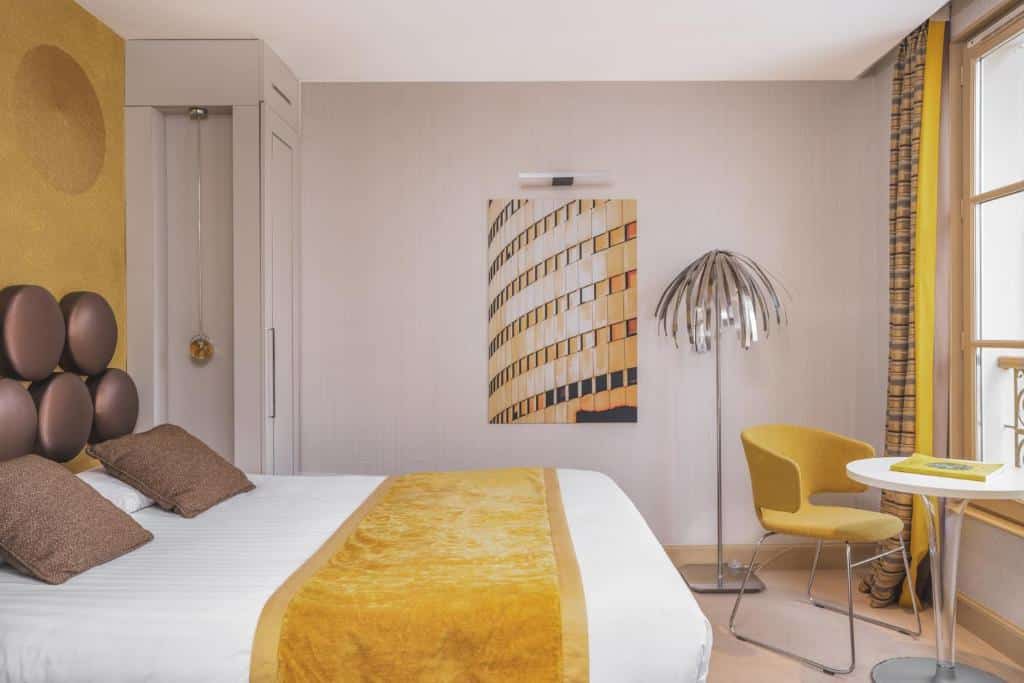 Quarto do Hotel Le petit Paris, um dos hotéis no Quartier Latin em Paris. Uma cama de casal está ao lado esquerdo da imagem, e uma mesa com abajur e cadeira fica na parede oposta, logo em frente a uma janela.