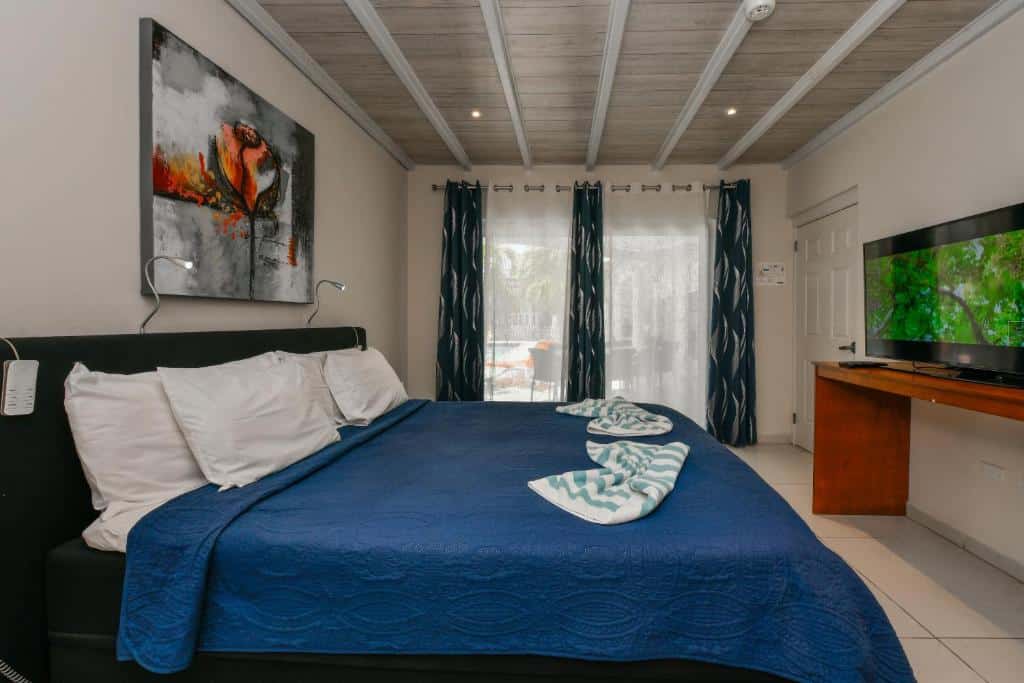 Quarto do Boutique Hotel Swiss Paradise Aruba Villas and Suites. Uma cama de casal está de frente ára uma grande tv. Ao lado da cama, mais ao fundo, está uma janela com cortinas.