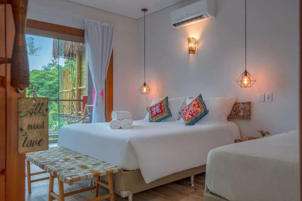 Quarto no hotel Vila Kebaya. Há uma cama de casal box no centro. Nos dois lados dela há uma luminária que está pendurada no teto. Acima da parede atrás do quarto há um ar-condicionado. Na esquerda da cama há uma porta de vidro com varanda.