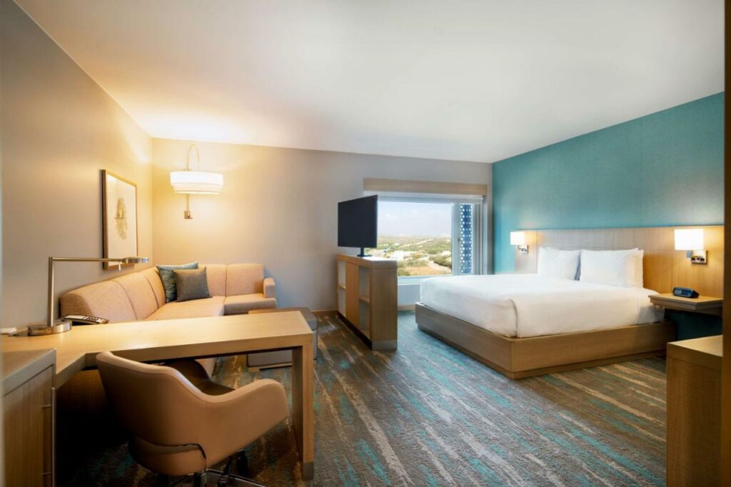 Quarto do Hyatt Place Aruba Airport com cama de casal no canto direito do espaço e uma área de sala de estar no canto esquerdo. A área conta com um grande sofá de canto bege, uma mesa de madeira com uma poltrona estofada e uma televisão de tela plana grande.