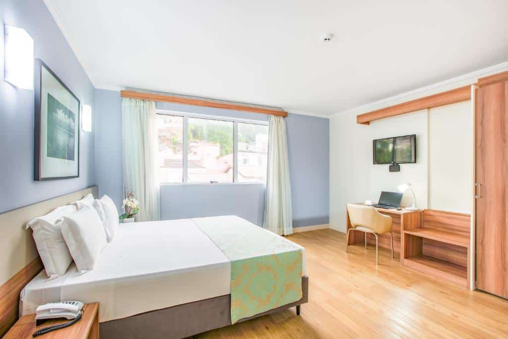 Foto do quarto do hotel Iate Hotel Centro Florianópolis. Ilustra o post sobre Onde ficar em Florianópolis. Na esquerda há uma cama box de casal, e vemos ela de lado. Na frente da cama há uma escrivaninha com cadeira, computador e uma TV, além de ter um armário ao lado. Do outro lado da cama há uma janela.