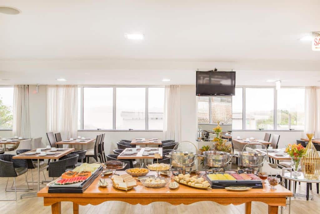 Foto mostrando o restaurante do Iate Hotel Centro Florianópolis enquanto o buffet de café da manhã é servido. A mesa do buffet está no centro, e atrás há outras mesas. No fundo tem várias janelas e uma TV.