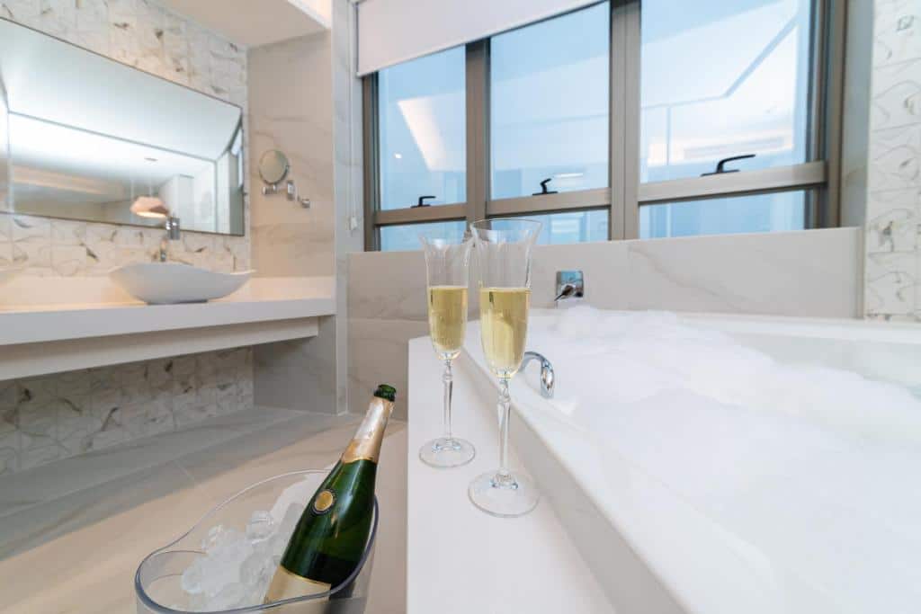 Um banheiro na Intercity Portofino Florianópolis. Há uma hidro na direita, e na beirada dela há duas taças de champagne. Fora da banheira há um balde com uma garrafa de champagne, perto da hidro. De frente da hidro há uma janela. No lado esquerdo tem uma pia e espelho.