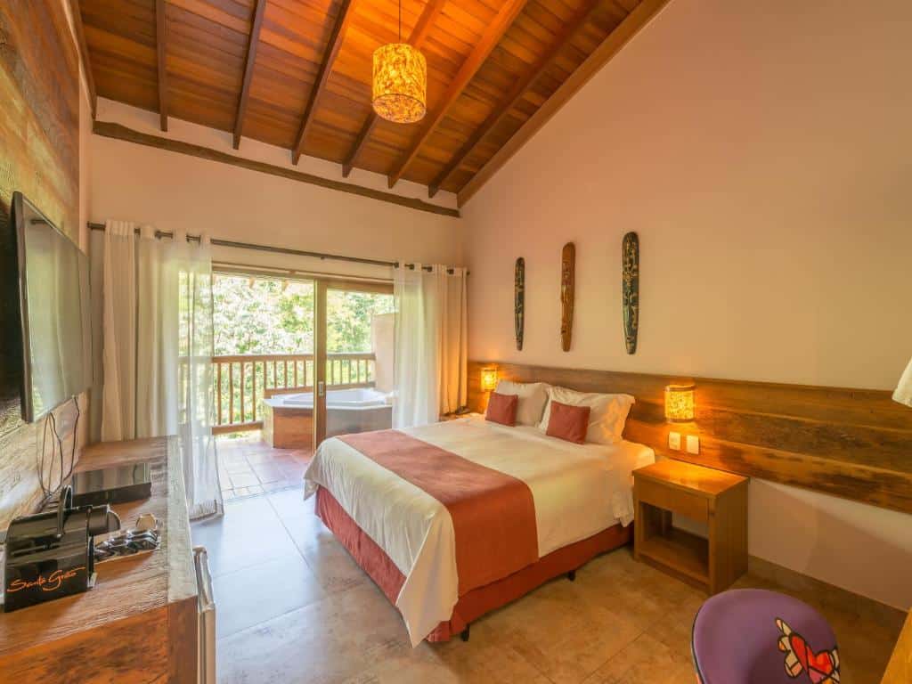 Foto do quarto em Kalango Hotel Boutique. Ilustra o post sobre Onde ficar em Ilhabela. Há uma cama box de casal no centro. Dos dois lados dela há uma mesa de cabeceira com luminária. Na frente da cama há um painel com televisão. No fundo há uma varanda com hidromassagem.