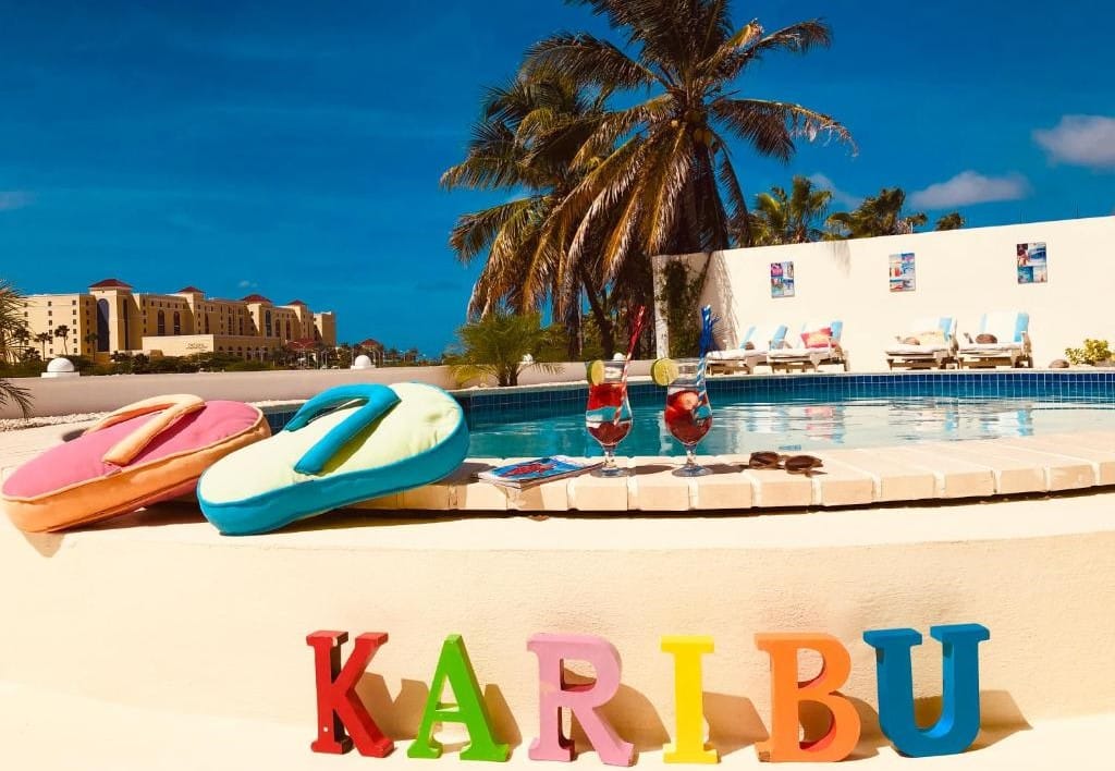 Piscina do Karibu Aruba Boutique Hotel. A foto foi tirada de baixo e motra o nome do hotel em frente a piscina, que possui duas taças e algumas almofadas em formato de chinelo na borda.
