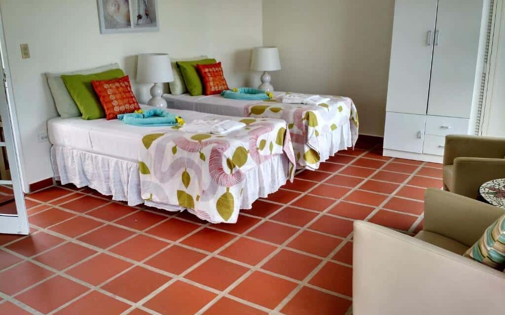 Quarto do hotel Karibu Aruba Boutique Hotel. Duas camas de solteiro estão lado a lado (com um abajur entre elas) do lado direito, ao fundo da imagem. Ao lado delas está um armário e em frente duas poltronas. Este é um dos hotéis baratos em Aruba.