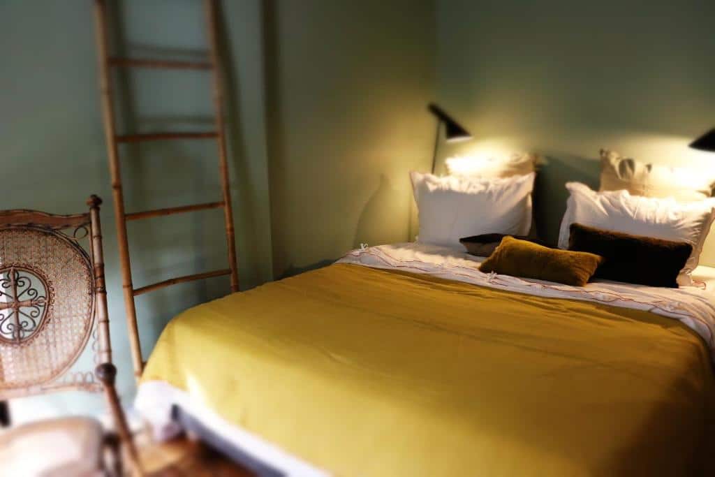Quarto do Le 66. Uma cama de casal está ao lado direito da imagem com luminárias dos dois lados e uma cadeira em frente. Na lateral da cama há uma escada de decoração.