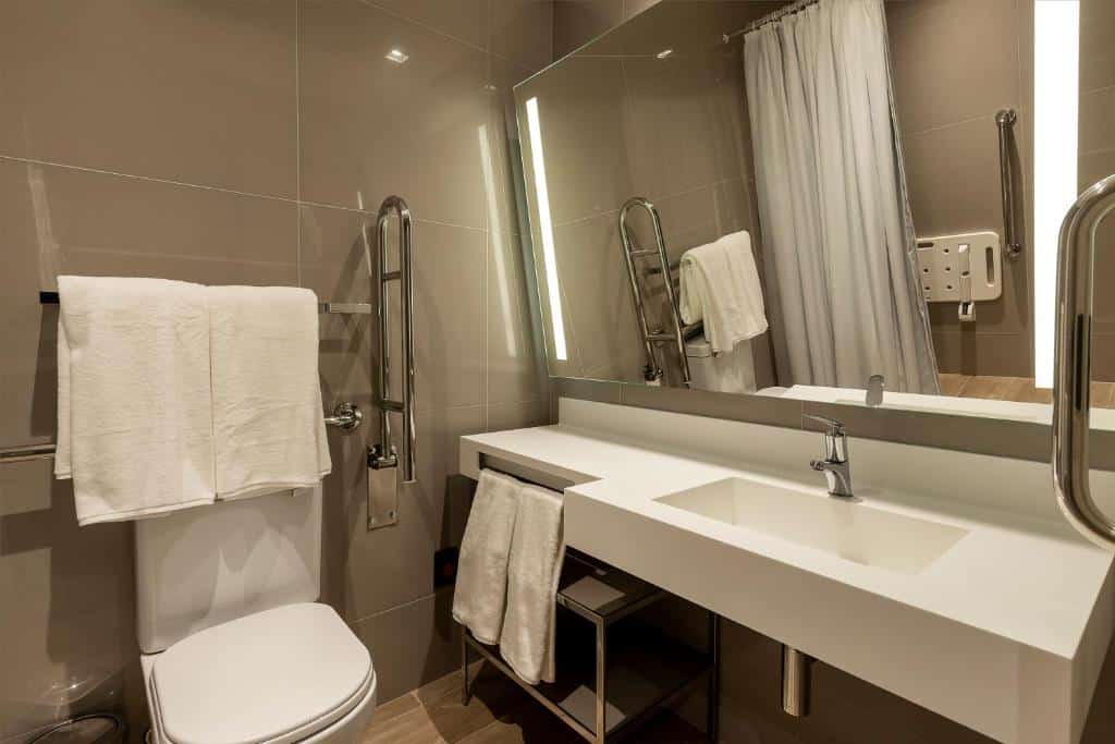 Banheiro moderno no LK Design Hotel Florianópolis. Há um vaso sanitário na esquerda e uma barra de apoio ao seu lado, na direita. Ocupando a direita da imagem vemos uma pia rebaixada com espelho inclinado para baixo. No reflexo do espelho vemos que há uma cadeira de banho na área da ducha.