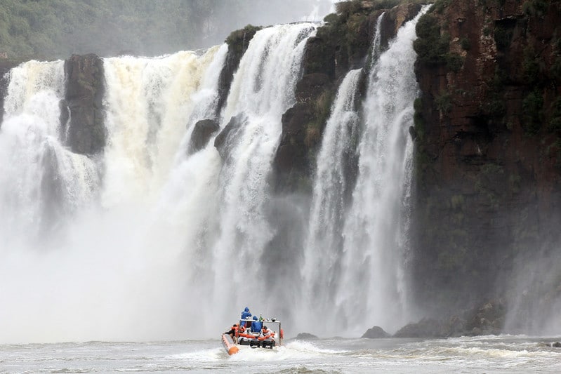 Foto mostrando um passeio em um bote no Rio Iguaçu, nas Cataratas. O bote está centralizado na imagem, e fica pequeno perto da queda d'água logo à sua frente.