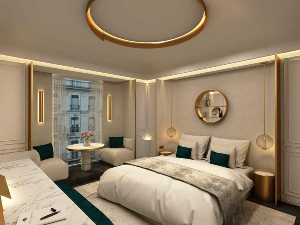 Quarto da Maison Albar - Le Vendome, um dos hotéis perto da Galeria Lafayette. Uma cama de casal está encostada na parede ao lado direito com mesinhas de cabeceira e luminárias dos dois lados, assim como uma decoração de parede logo acima. A lateral esquerda da cama tem uma janelas com cortinas, e uma mesinha com duas poltronas fica logo em frente à janela.