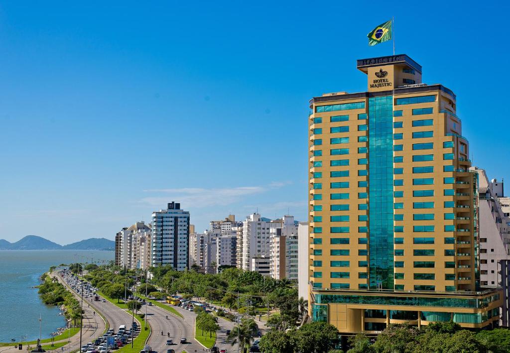 Vista do exterior do Majestic Palace Hotel. O hotel em questão aparece mais na direita, e há outros hotéis ao fundo. Na direita vemos algumas avenidas beira-mar no centro de Florianópolis. O céu está bem azul.