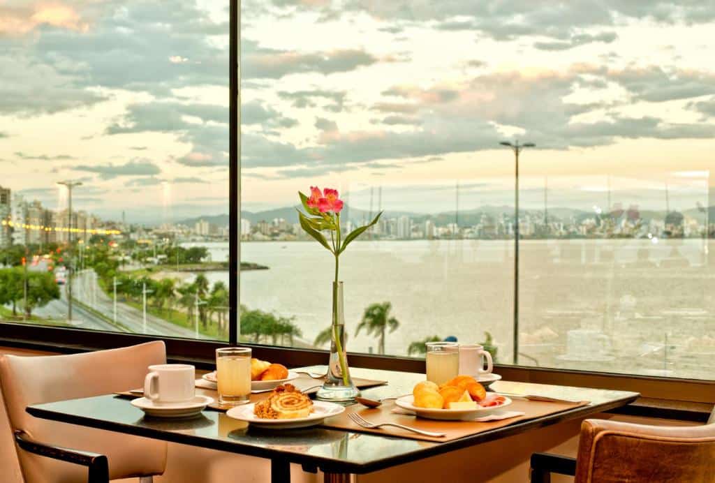Um exemplo de café da manhã no Majestic Palace Hotel. Mostra uma mesa com xícaras, copos com suco, doces, etc. No centro da mesa tem um vaso de vidro com flor. A vista do lado da mesa é para uma praia de Florianópolis.