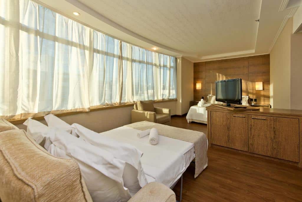 Foto do quarto do hotel Majestic Palace Hotel. Ilustra o post sobre Onde ficar em Florianópolis. Há uma cama na esquerda, e na frente dela há uma estante com TV e uma poltrona. Do lado esquerdo da cama há uma janela que se estende até o fundo do quarto.