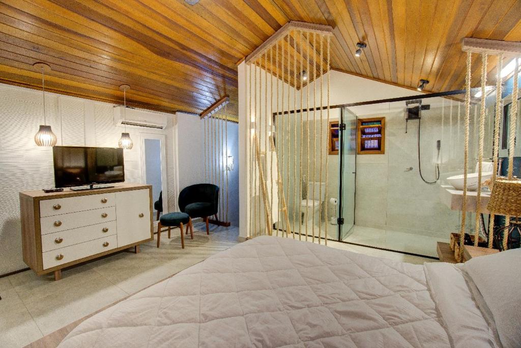 Quarto do Malie Chalés. Uma cama de casal e no fundo o box e banheiro de vidro. Do lado esquerdo uma cômoda, uma poltrona, um espelho a tv e o ar-condicionado.