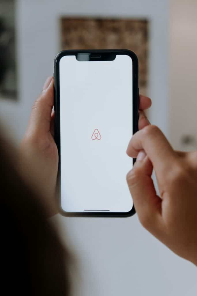 Mãos brancas segurando um celular com a tela branca e o logo do airbnb no meio.O fundo está embaçado