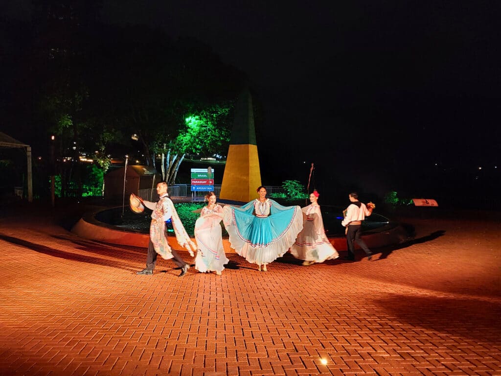 Foto do Show no Marco das Três Fronteiras do lado brasileiro. Há três mulheres com roupas características e também dois homens. Foto tirada à noite em um pátio.