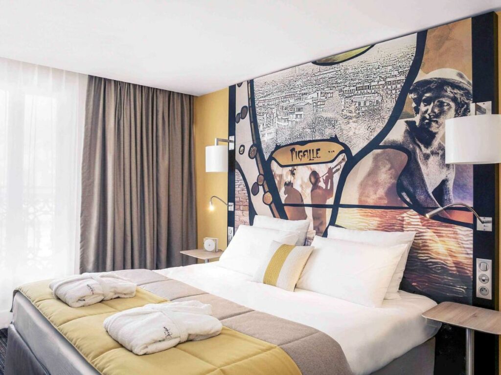 Quarto do Mercure Paris Pigalle Sacre Coeur. Uma cama de casal está encostada na parede ao lado direito com mesinhas de cabeceira e luminárias aos dois lados, assim como roupões dobrados aos pés. Ao lado fica uma janela com cortinas.