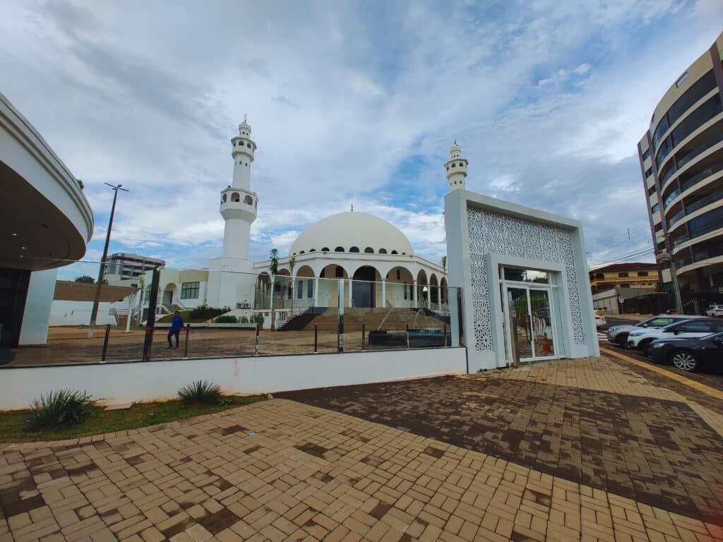 A mesquita Omar Ibn Al-Khattab tem uma cúpula branca grande e duas torres (minaretes) ao lado. A entrada é marcada por um portão decorado com padrões islâmicos, e ao redor há um pavimento de tijolos.