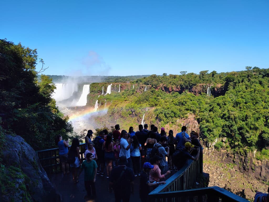 Foto de várias pessoas agrupadas em um mirante para as cataratas do Iguaçu. As cataratas estão no fundo direito, e tem um arco-íris de frente para elas.