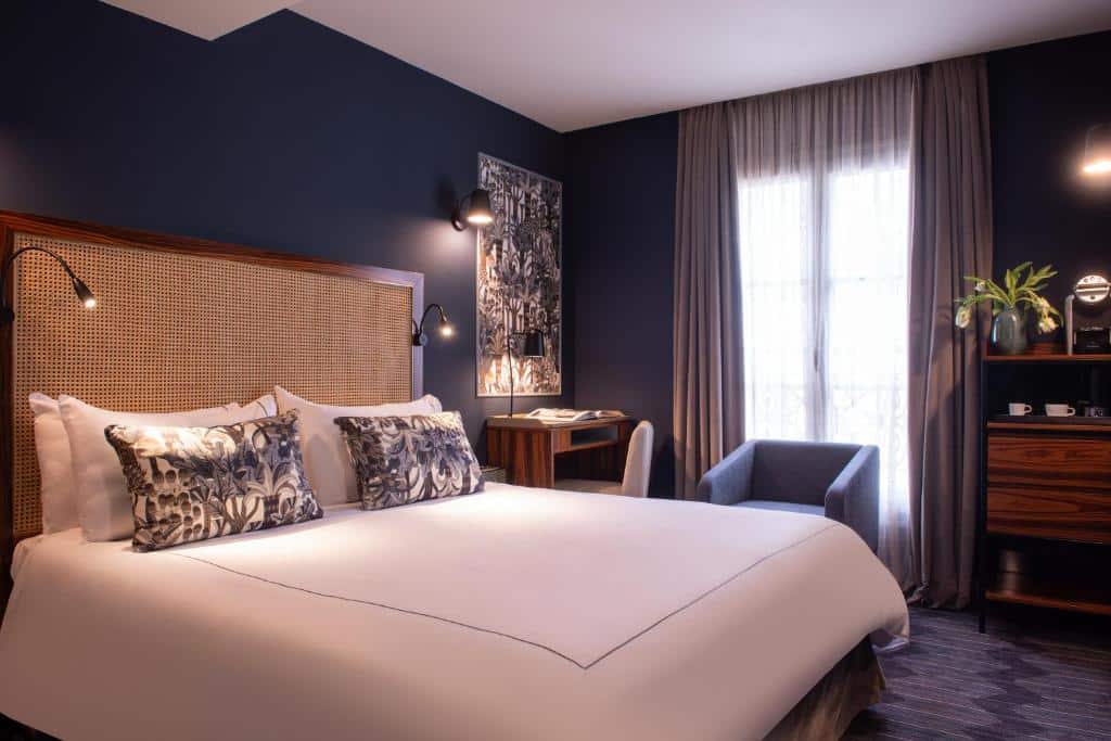 Quarto do Mom'Art Hotel, um dos hotéis 4 estrelas em Paris. Uma cama de casal está encostada na parede ao lado esquerdo com mesinhas de cabeceira e luminárias aos dois lados. Ao lado fica uma mesinha com cadeira e luminária, e atrás há uma poltrona. Há uma janela atrás da poltrona, e ao lado fica uma cômoda.