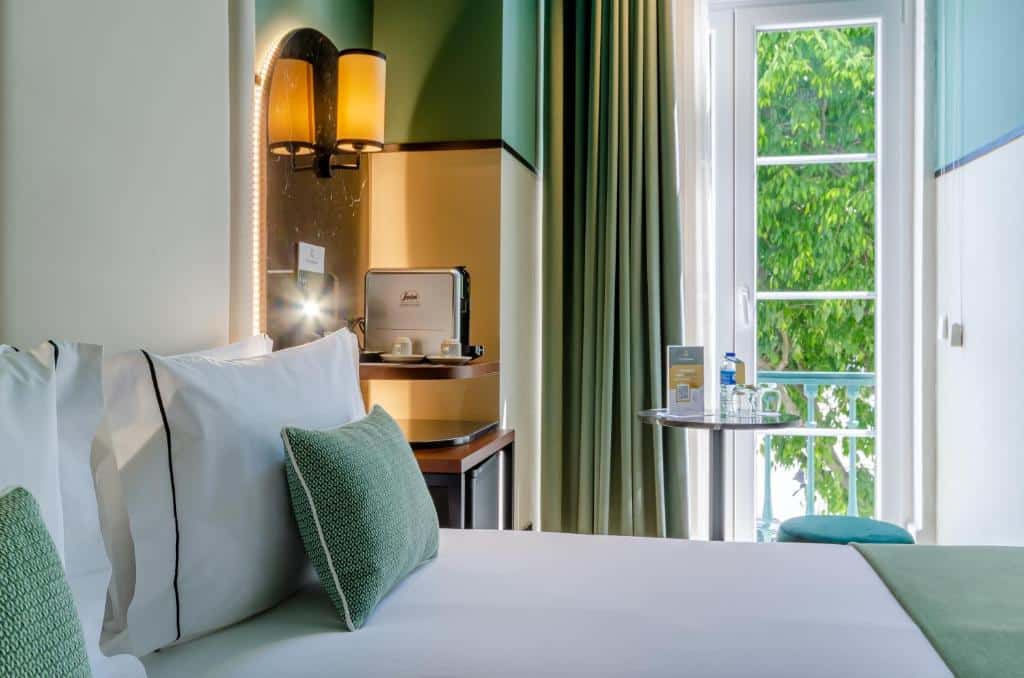 Quarto do Nicola Rossio Hotel com uma cama de casal e, ao lado dela, há uma sacada de vidro com vista para as árvores da Praça do Rossio, além de comodidades para preparar café, um frigobar e uma pequena mesa com um pufê redondo