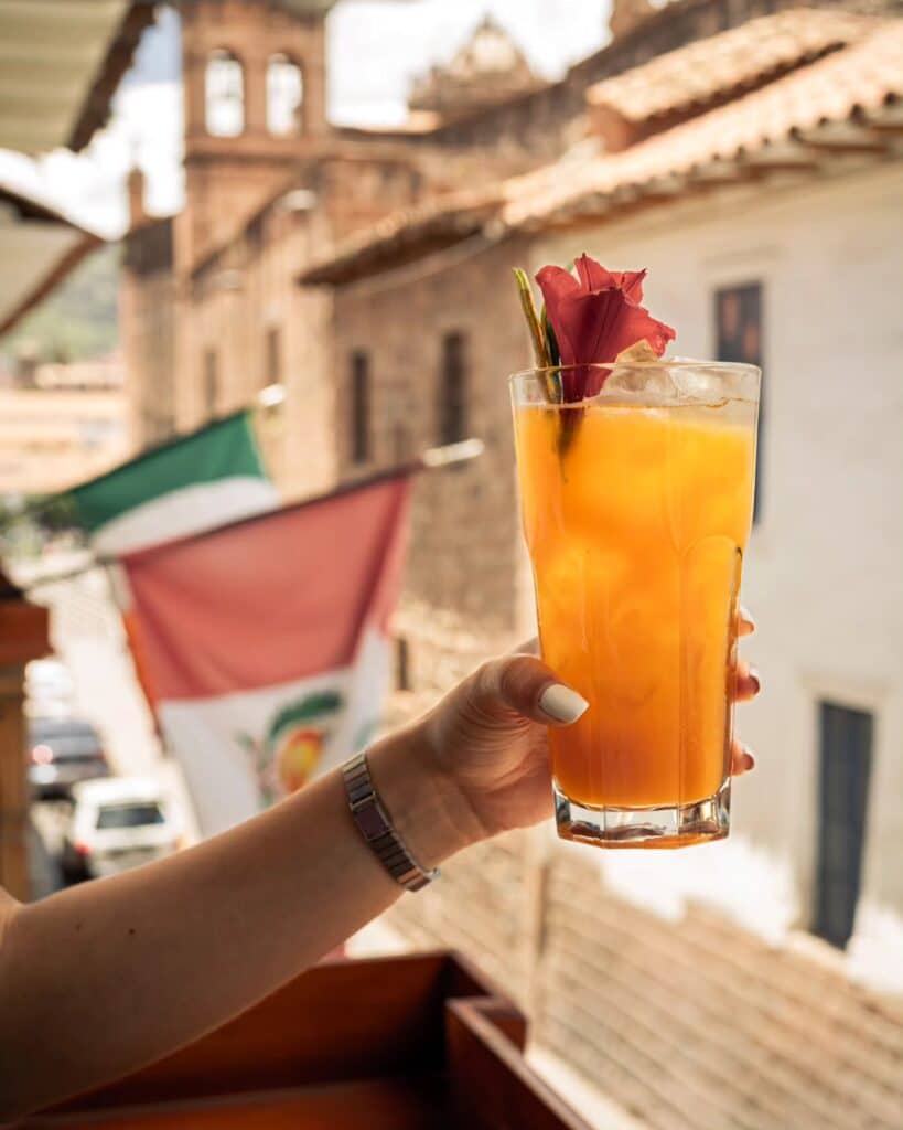 um braço feminino branco segurando um copo de bebida refrescante com a praça de armas em Cusco aparecendo ao fundo