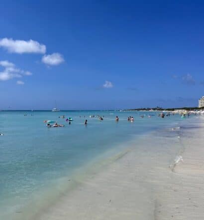 Imagem de Palm Beach para ilustrar post sobre onde ficar em Aruba. Um mar cristalino do lado esquerdo com algumas pessoas, do lado direito a areia e pessoas caminhando.