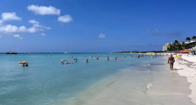 Imagem de Palm Beach para ilustrar post sobre onde ficar em Aruba. Um mar cristalino do lado esquerdo com algumas pessoas, do lado direito a areia e pessoas caminhando.
