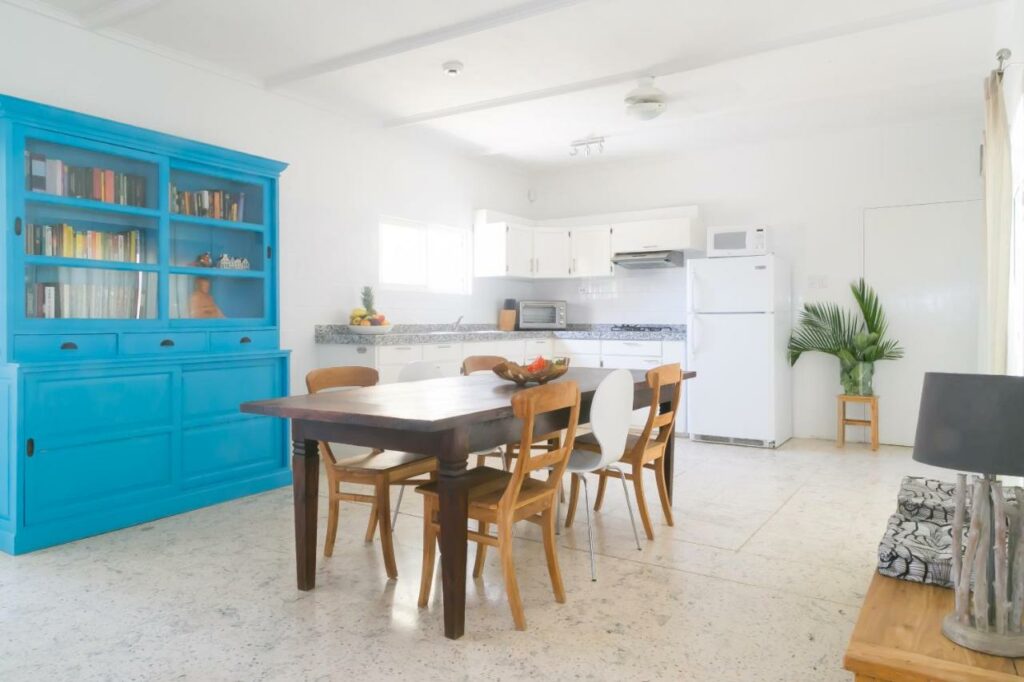 Cozinha do Pauline’s Apartments. Uma mesa de refeições está no centro da imagem. Na parede esquerda estão um armário, a pia, a geladeira e outros eletrodomésticos. Foto para ilustrar o post de hotéis baratos em Aruba.