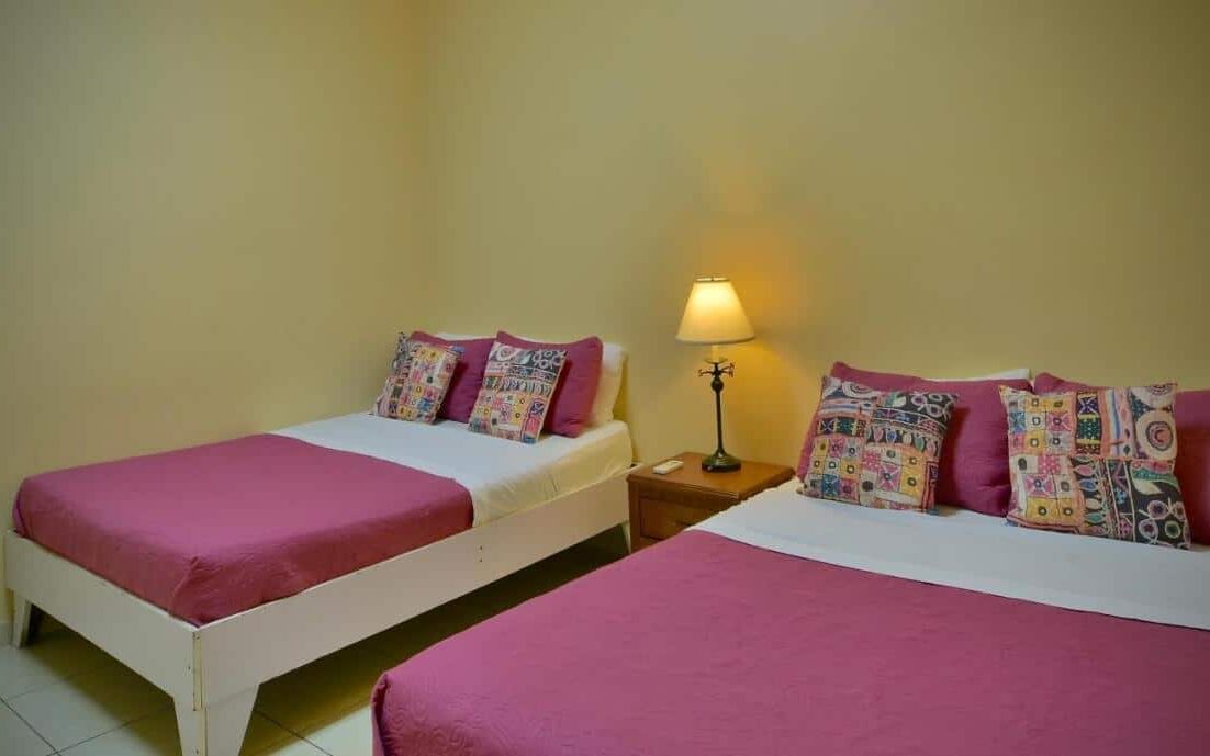 Um dos quartos compartilhados do Pista Q hostel and apartments. Uma cama de casal e uma de solteiro estão lado a lado com uma mesinha com abajur entre elas.