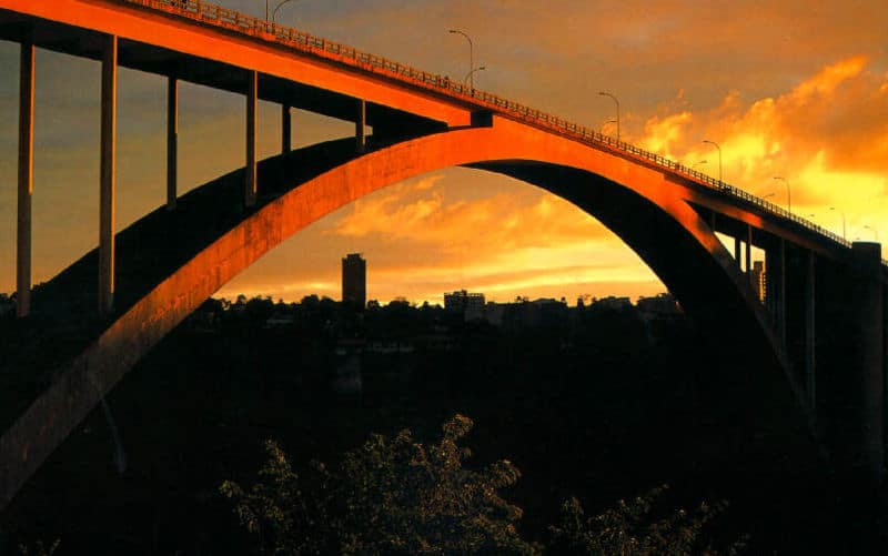 Foto tirada durante um pôr do sol que mostra a Ponte Internacional da Amizade. O céu está alaranjado e possui algumas nuvens.