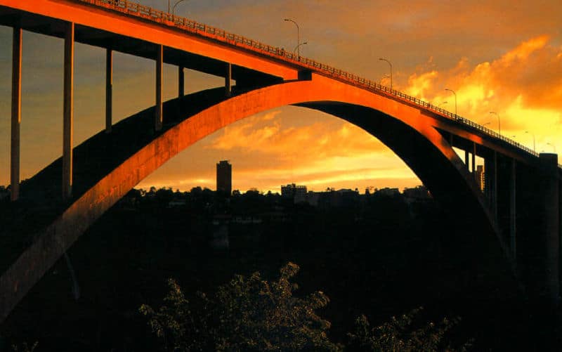 Foto tirada durante um pôr do sol que mostra a Ponte Internacional da Amizade. O céu está alaranjado e possui algumas nuvens. 