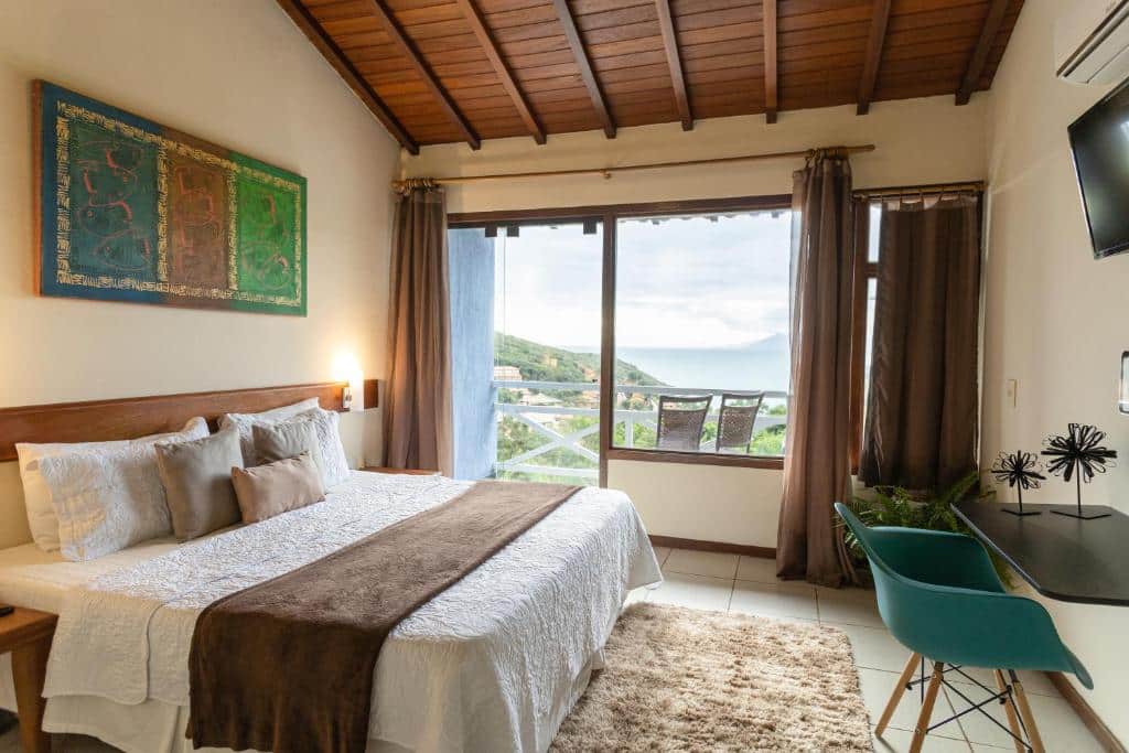 Quarto da Pousada Amancay. Do lado esquerdo a cama, de frente uma mesa com cadeira, em cima um ventilador. No fundo a porta da varanda e vista do mar.