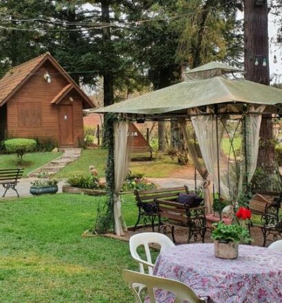 Área externa da Pousada Árvore Da Coruja para ilustrar post sobre pousadas pet friendly em Gramado. Uma mesa com quatro cadeiras do lado direito, atrás uma tenda com mesas e cadeiras de ferro, no fundo os chalés.