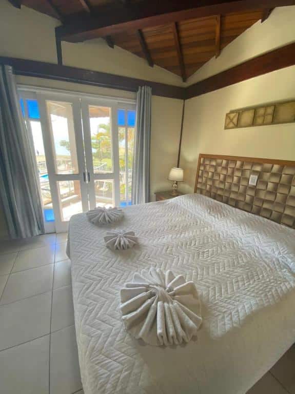 Foto do quarto em Pousada Bonns Ventos. Ilustra o post sobre Onde ficar em Ilhabela. Há uma cama box de casal na direita, e do lado da cama há uma porta com varanda e vista.