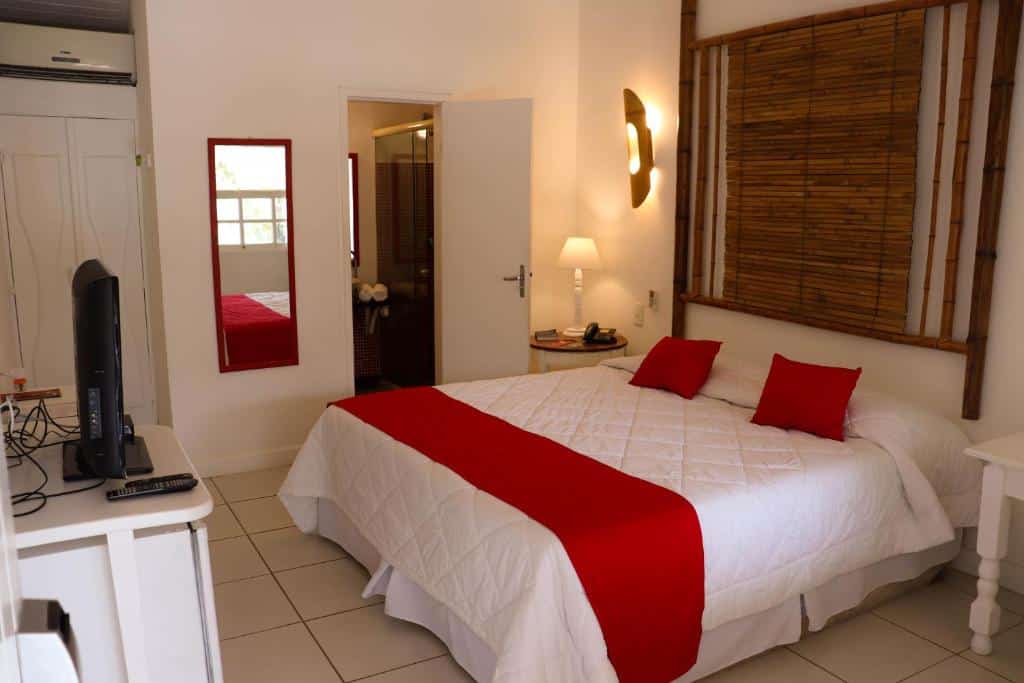 Quarto da Pousada Bucaneiro. Uma cama de casal do lado direito com uma mesinha de apoio de cada lado com luminária. Do lado esquerdo uma mesa com televisão, em baixo o frigobar. No fundo um ar-condicionado, um espelho e a porta do banheiro. Foto para ilustrar post sobre pousadas pet friendly em Búzios.