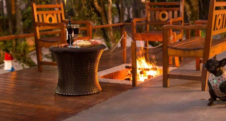 A imagem mostra uma área de convivência da Pousada Campos dos Holandeses, em que há no centro uma fogueira saindo do chão, cercada por bancos e cadeiras, além de uma mesa com taças de vinho e uma tábua de frios. No lado direito da imagem é possível ver um cachorro da raça Pug, usando um coletinho. Imagem utilizada para ilustrar o post pousadas pet friendly em Campos do Jordão