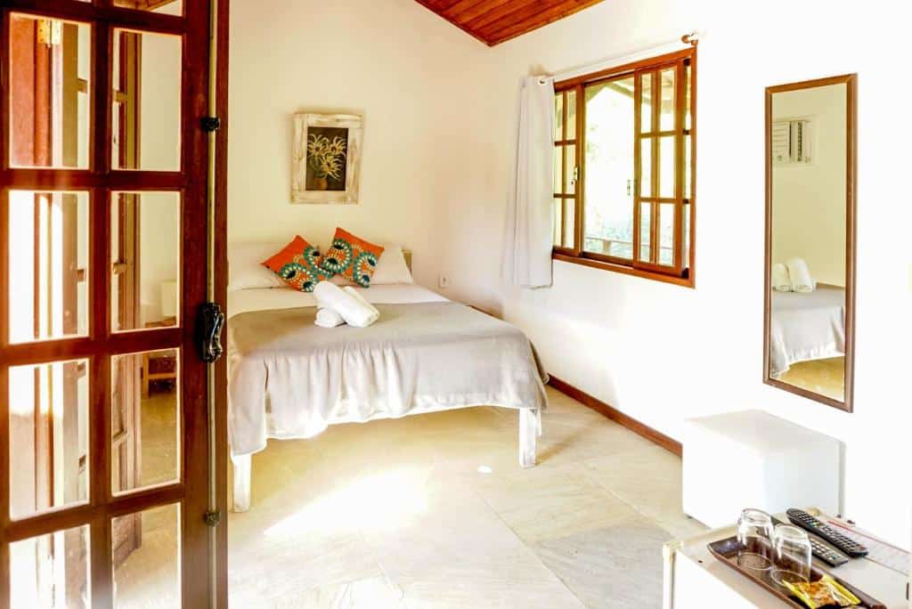 Quarto da Pousada Casa Cactus. Uma cama de casal no fundo, do lado direito uma janela, um espelho e um puff. Na ponta da direita o frigobar com copos e controles de TV. Foto para ilustrar post sobre pousadas pet friendly em Búzios.