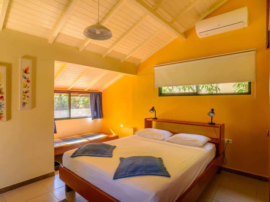 Foto do quarto em Pousada Ecoilha. Ilustra o post sobre Onde ficar em Ilhabela. Há uma cama de casal no centro. Do lado esquerdo da cama há um banco e uma janela. Atrás da cama há outra janela. No topo da parede à direita há um ar-condicionado.