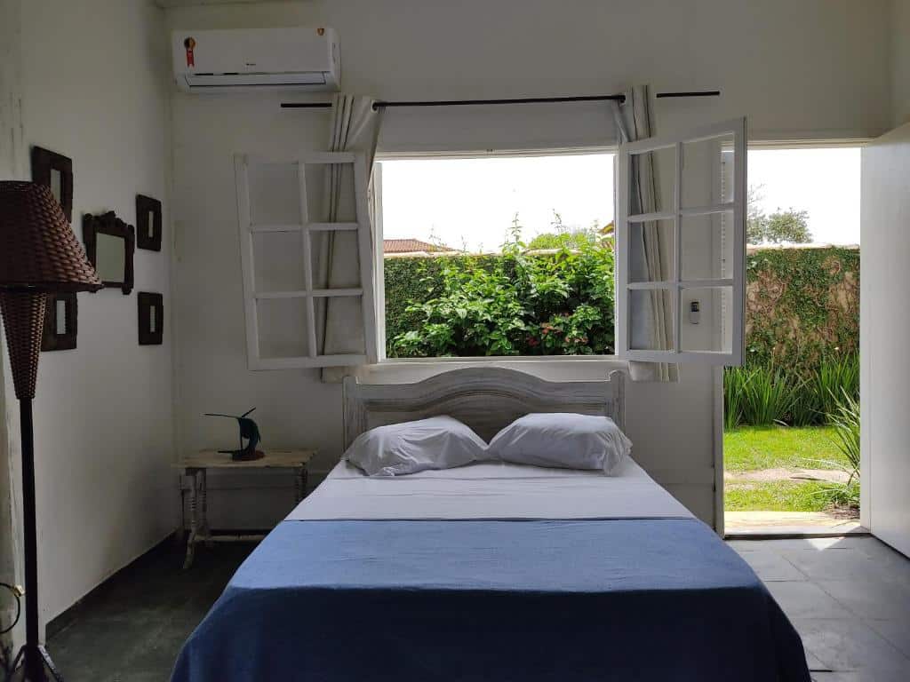 Quarto da Pousada Manaaki. Uma cama de casal no meio, do lado esquerdo um abajur e uma mesa de apoio. Em cima da cama uma janela, do lado direito a porta do quarto. Foto para ilustrar post sobre pousadas pet friendly em Búzios.