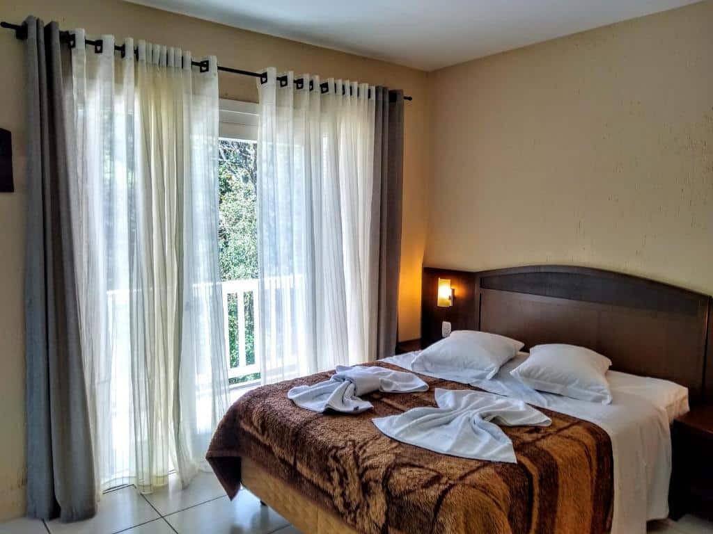 Quarto da Pousada Nosso Lar. Uma cama de casal com cobertor e toalhas, no canto uma luminária. No fundo a varanda com cortinas.