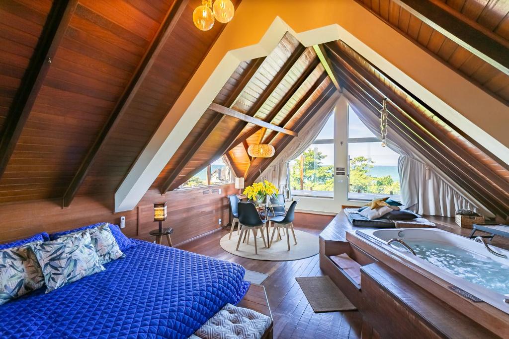 Foto do quarto do hotel Pousada Penareia Floripa. Ilustra o post sobre Onde ficar em Florianópolis. Há uma cama na esquerda e uma hidro na direita. O teto é em formato de triângulo. No fundo há uma mesa com cadeiras e uma janela com vista para o mar.