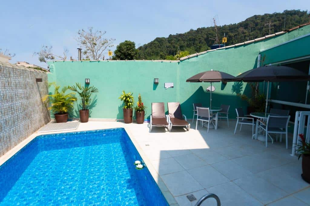 Área da piscina da Pousada Pérola de Ubatuba com uma piscina do lado esquerdo e algumas cadeiras com guarda-sóis do lado direito