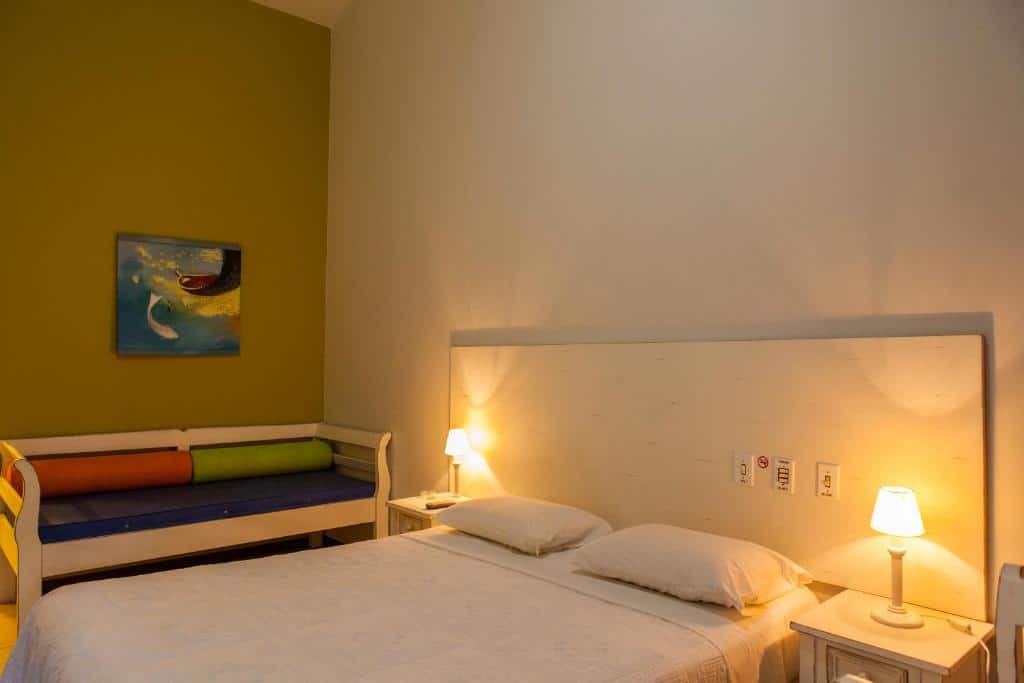 Quarto da Pousada Ponta do Lobo. Uma cama de casal com uma cômoda e um abajur de cada lado, no fundo um sofá cama. Foto para ilustrar post sobre pousadas pet friedly em Santa Catarina.