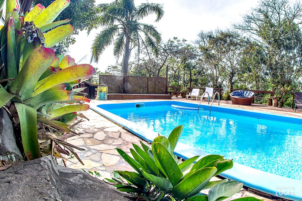 Área externa da Pousada Rosa. Umas plantas do lado esquerdo, do lado direito uma piscina com cadeiras de tomar sol, sofá e árvores.