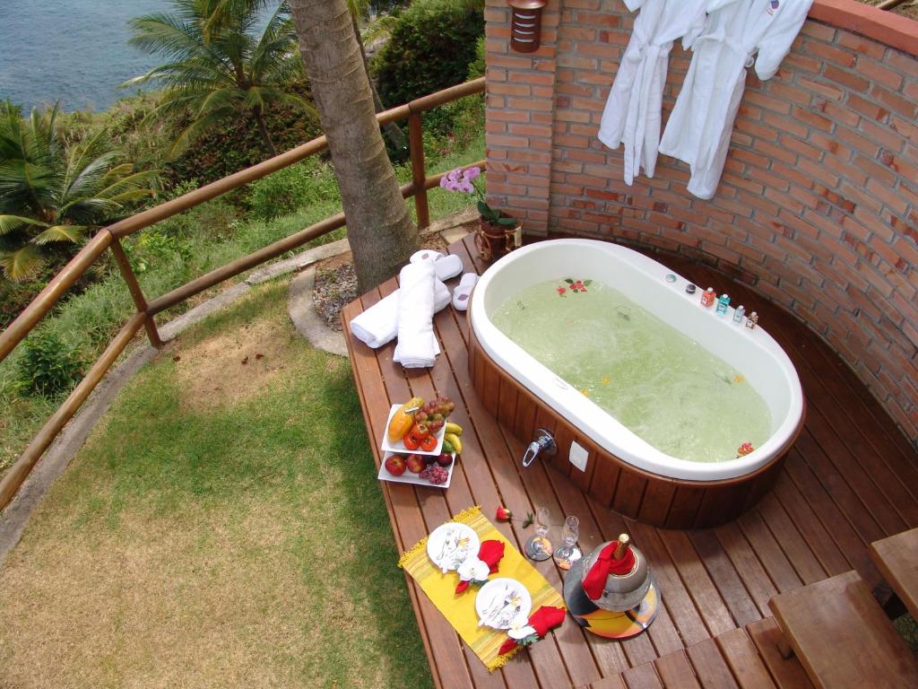 Foto aérea tirada de um jardim com um deck na Pousada Singuitta que contém uma hidro ao ar livre. E do lado da hidro há várias frutas, um balde de champagne, e dois pratos com talheres.