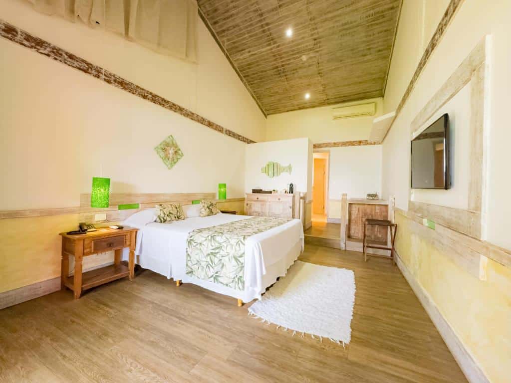 Foto do quarto em Pousada Vila das Velas. Ilustra o post sobre Onde ficar em Ilhabela. Há uma cama no centro, encostada na parede à esquerda. Na frente dela, na direita, há uma televisão. No fundo do quarto há uma divisória e uma porta. O quarto é bastante espaçoso.