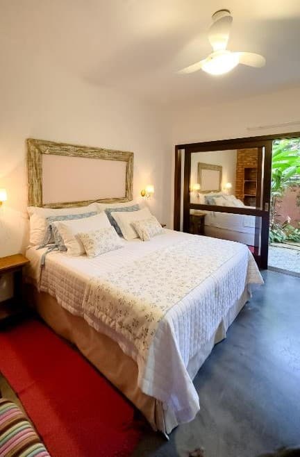 Quarto da Pousada Vila Rosada. Uma cama de casal, de cada lado uma mesinha de apoio e uma luminária. No fundo a porta da varanda. Foto para ilustrar post sobre pousadas pet friedly em Santa Catarina.